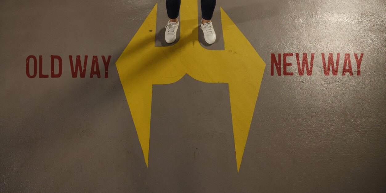 Bild von Füßen, die auf einem auf den Boden aufgemalten Pfeil stehen, der in zwei Richtungen zeigt, mit Beschriftung "old way" und "new way"